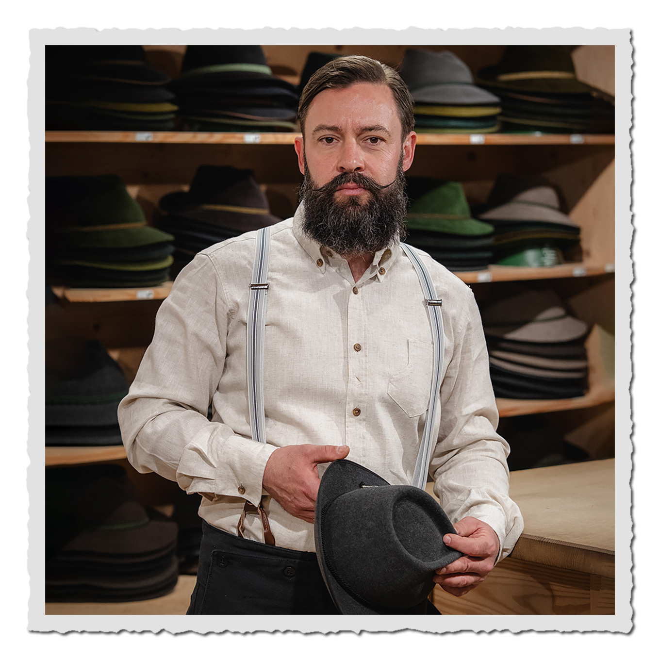 Mann mit Bart in Hutwerkstatt, trägt helles Hemd mit Hosenträgern und hält einen grauen Filzhut.